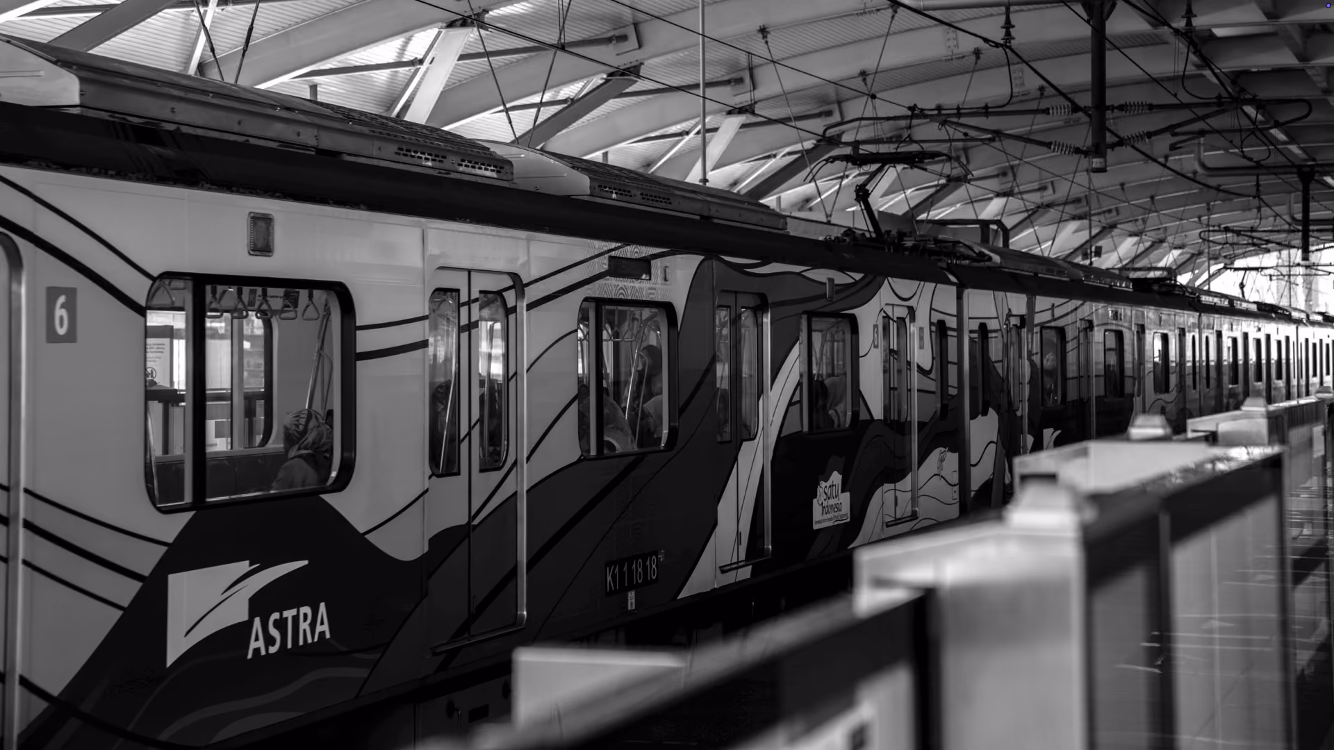 Black and white photo of a train at a station platform with the word 'ASTRA' visible on the side, under a high roof structure with passengers seated inside.