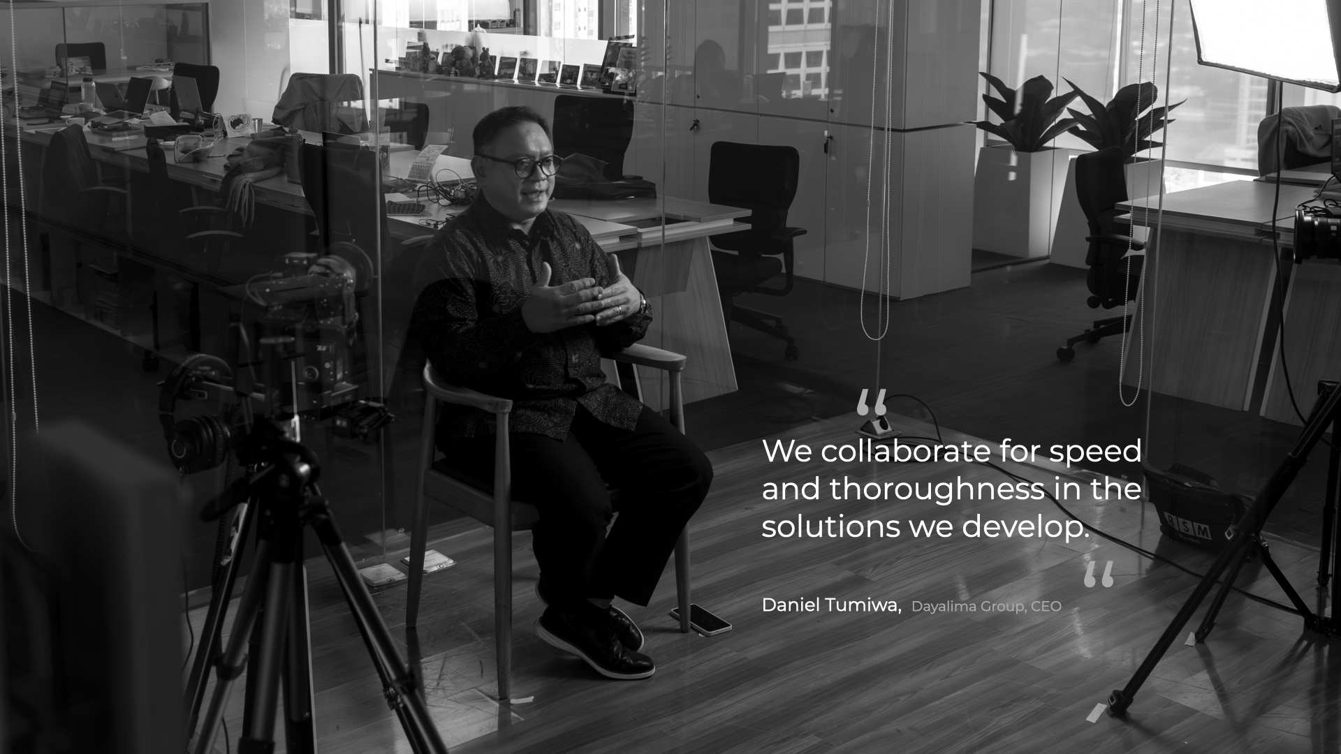 Man wearing glasses and a patterned shirt sitting on a wooden chair in an office, surrounded by cameras and lighting equipment, speaking with hands gesturing. A quote overlay reads, 'We collaborate for speed and thoroughness in the solutions we develop.' attributed to Daniel Tumiwa, Dayalima Group, CEO.