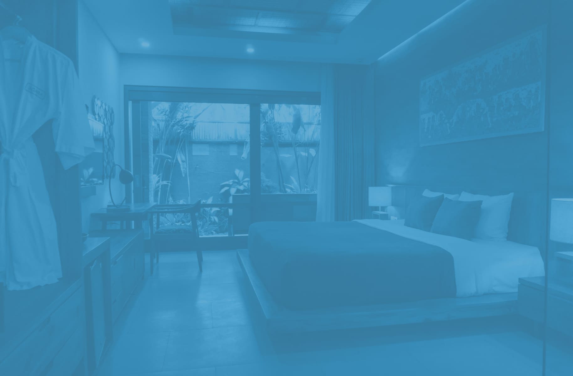 Modern bedroom with a large bed, bedside lamps, desk, chair, and a window showing outdoor plants.