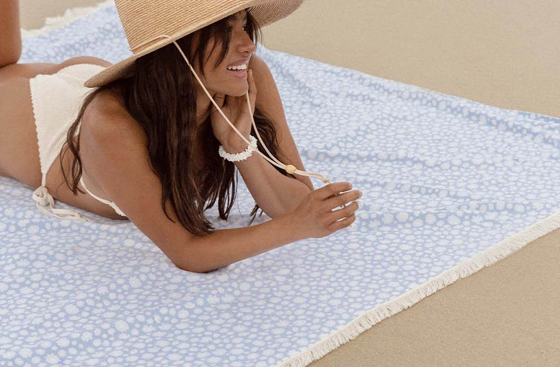 Woman in a white bikini and sun hat lying on a blue patterned beach towel on sand.