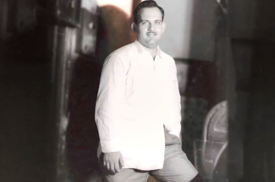 Black and white photo of a smiling man in a light shirt leaning against furniture in a dim room.
