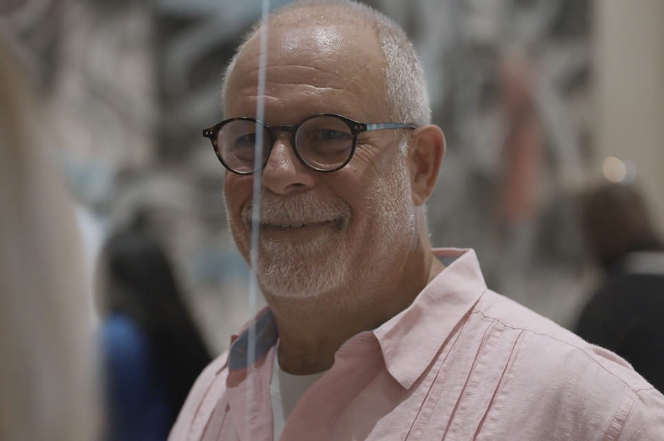 Smiling older man with glasses and grey beard wearing a light pink shirt in an indoor setting.