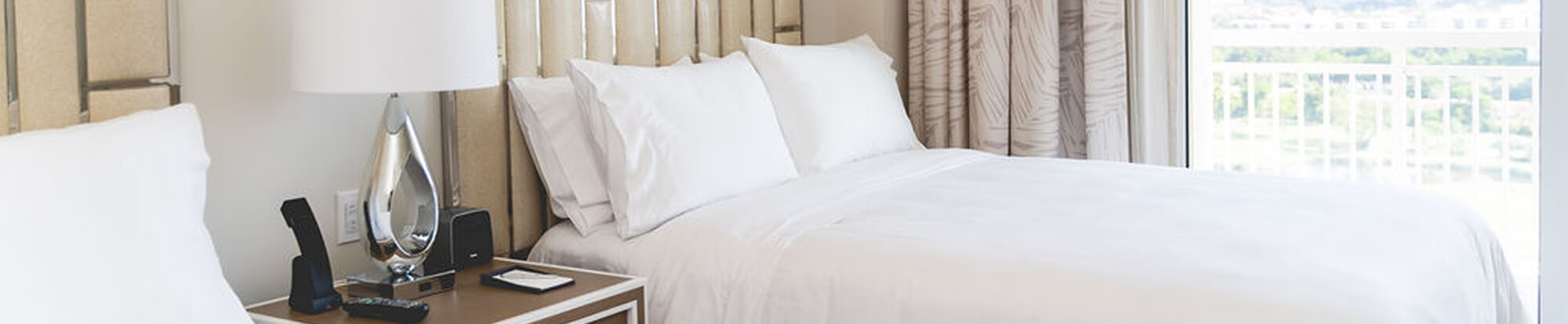 Bright hotel room with white bedding, pillows, bedside lamp, and balcony with railing outside.