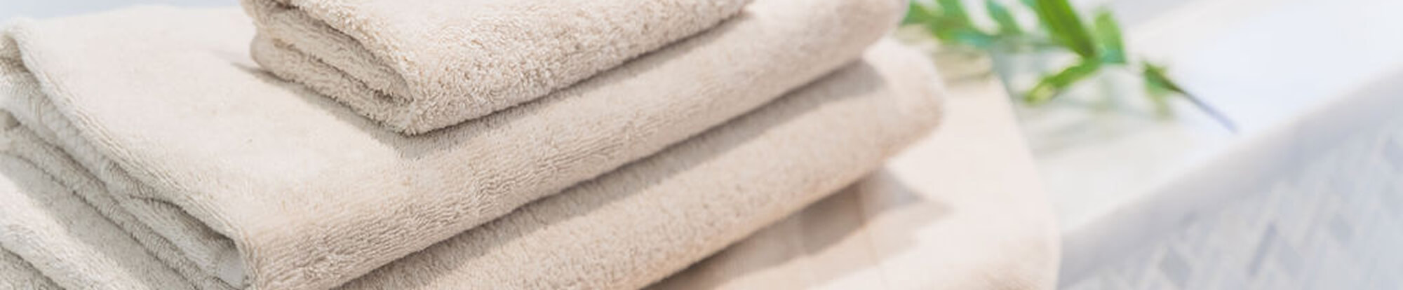 Stack of beige and cream folded towels on a light surface with a blurred green plant in the background.