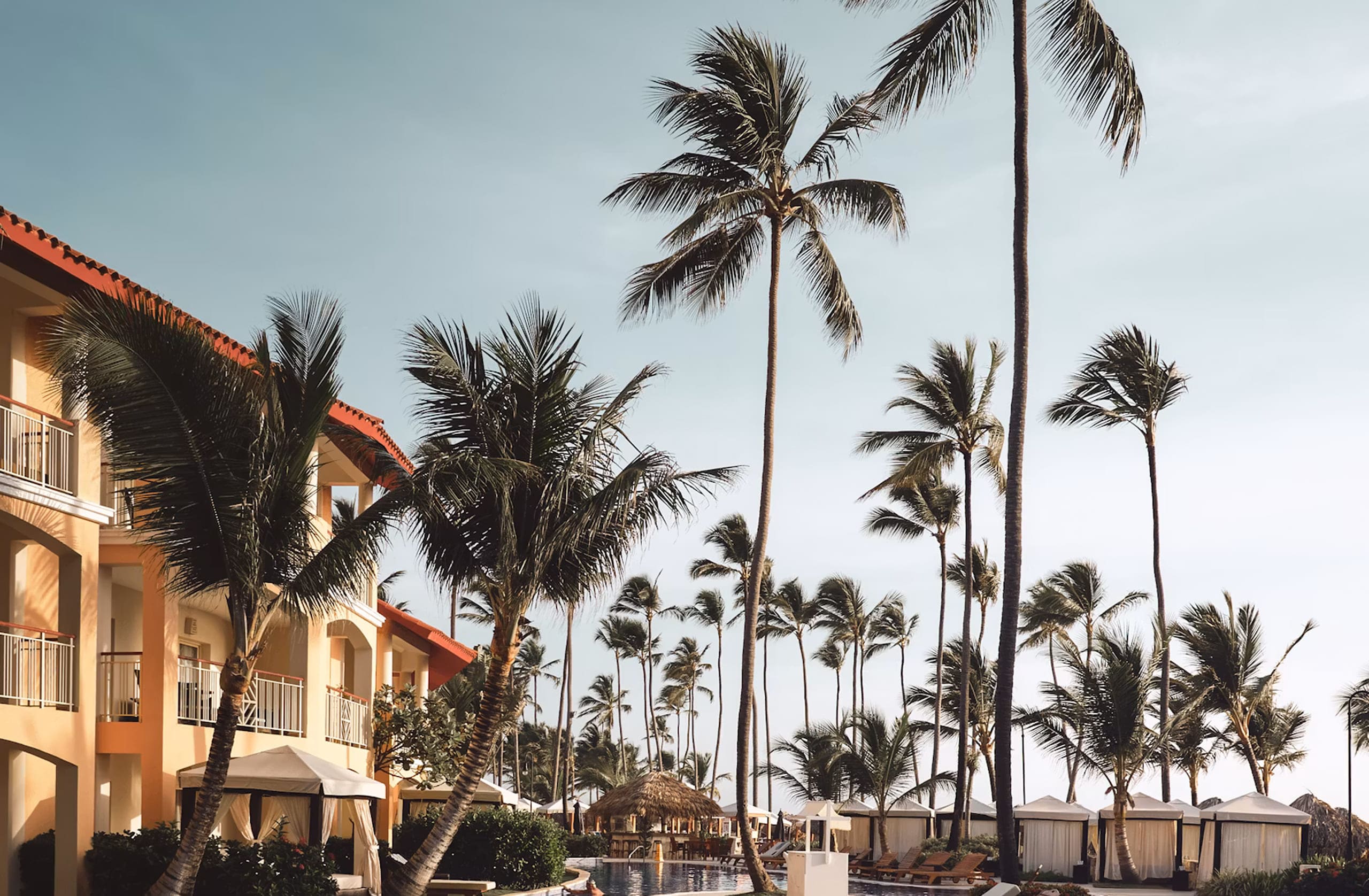 Palm trees around a resort pool with cabanas and lounge chairs under a clear sky.
