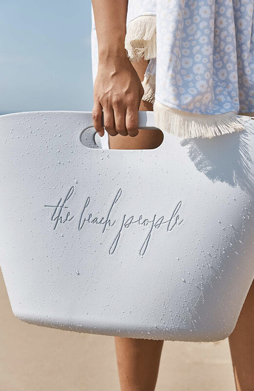 Person holding a light blue textured bag with ‘the beach people’ text near sandy shore.