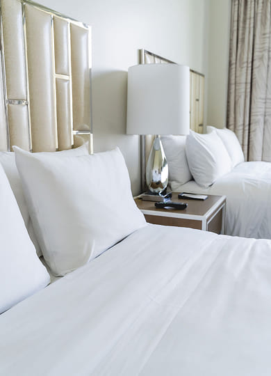 Two neatly made beds with white linens and pillows, separated by a nightstand with a silver lamp and remote controls.