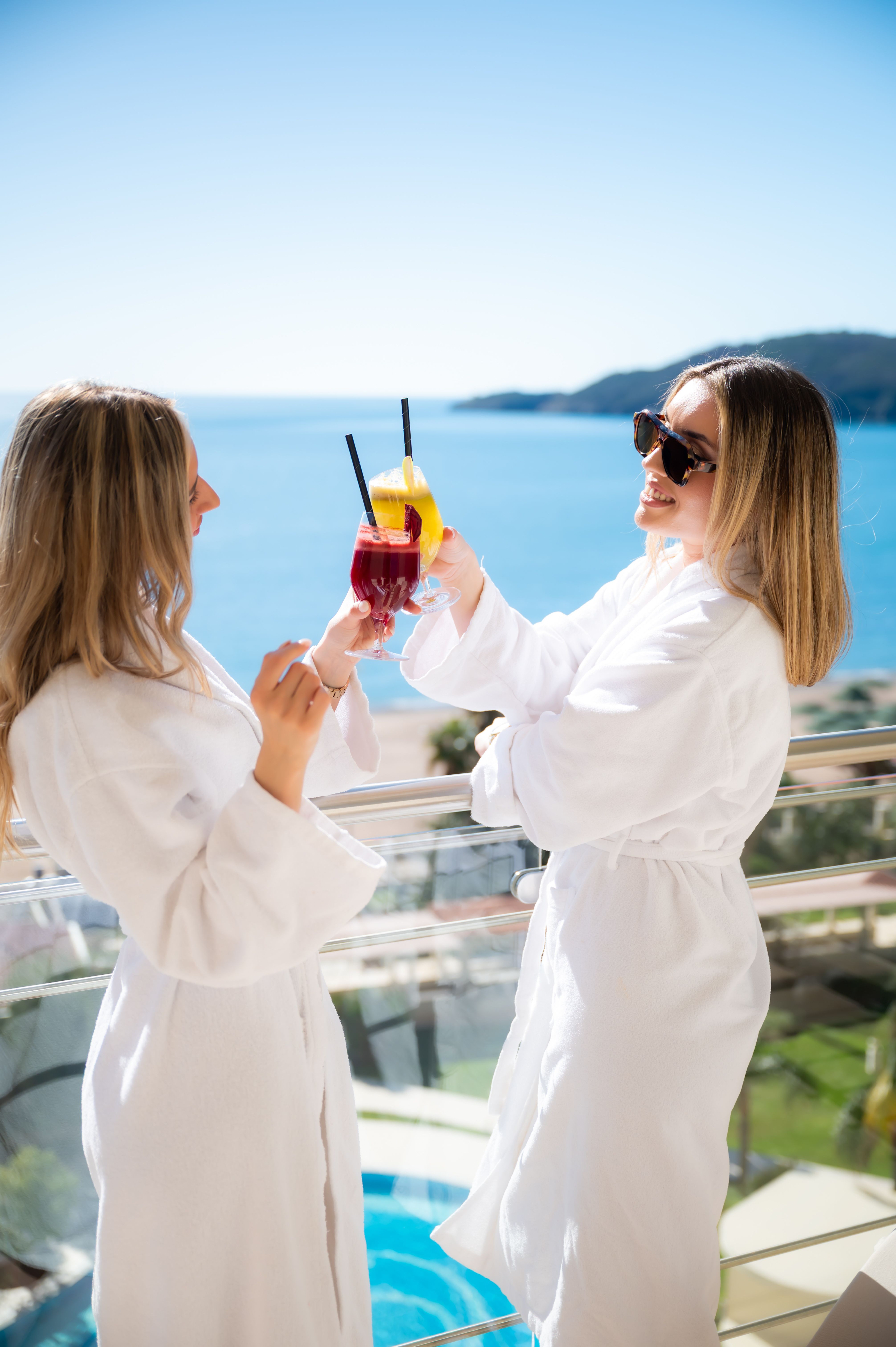 Two women in white robes toasting colorful drinks on a balcony overlooking the sea and hills.