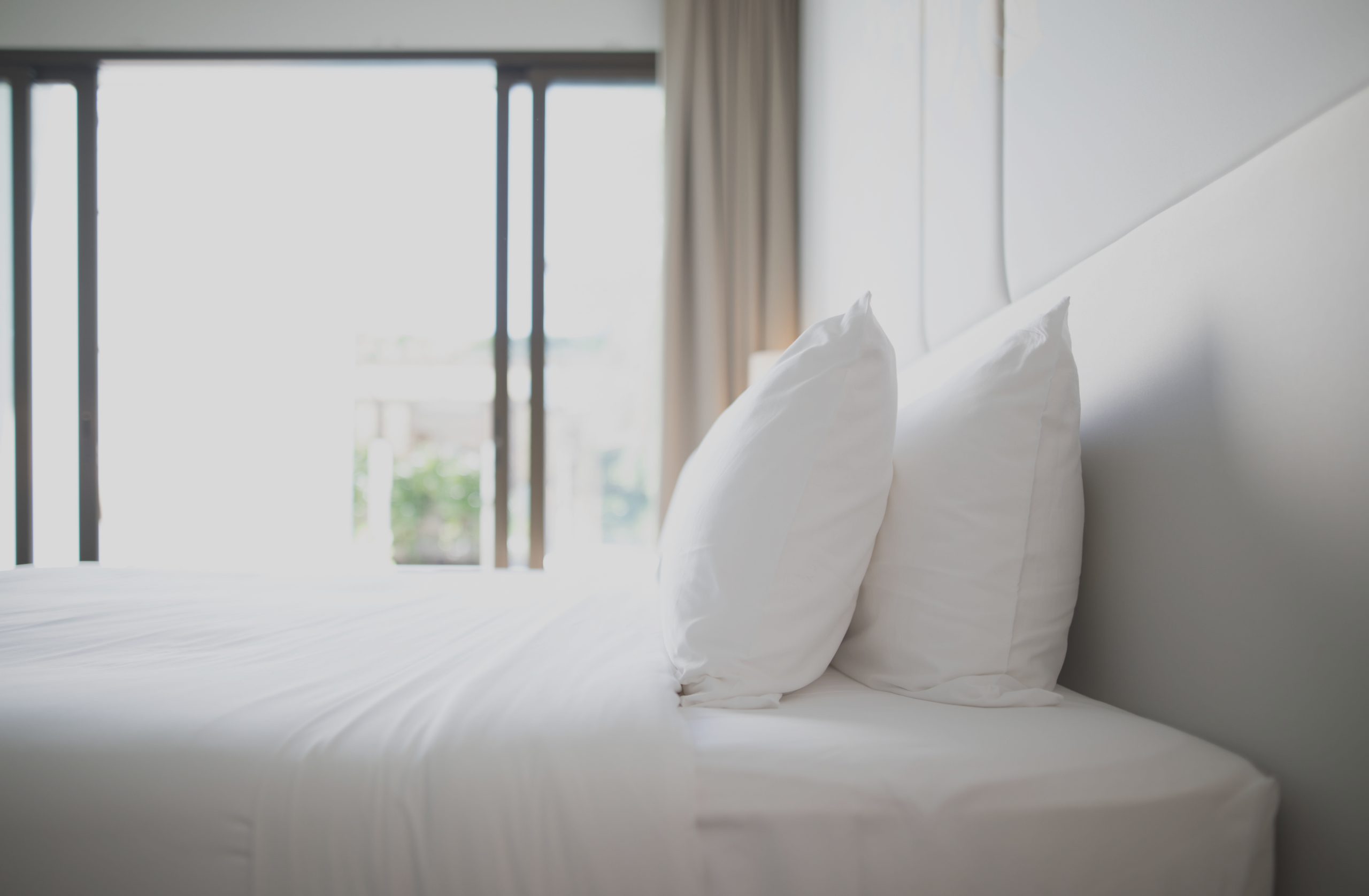Bright bedroom with a neatly made white bed and two pillows near a large window with curtains.