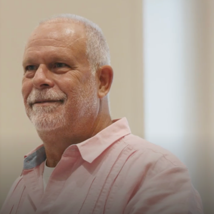 Portrait of a smiling middle-aged man with short gray hair and a beard wearing a light pink shirt.