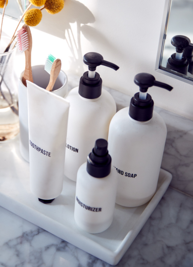 Toothpaste tube, wooden toothbrushes, lotion, hand soap, and moisturizer bottles on marble tray by sink.