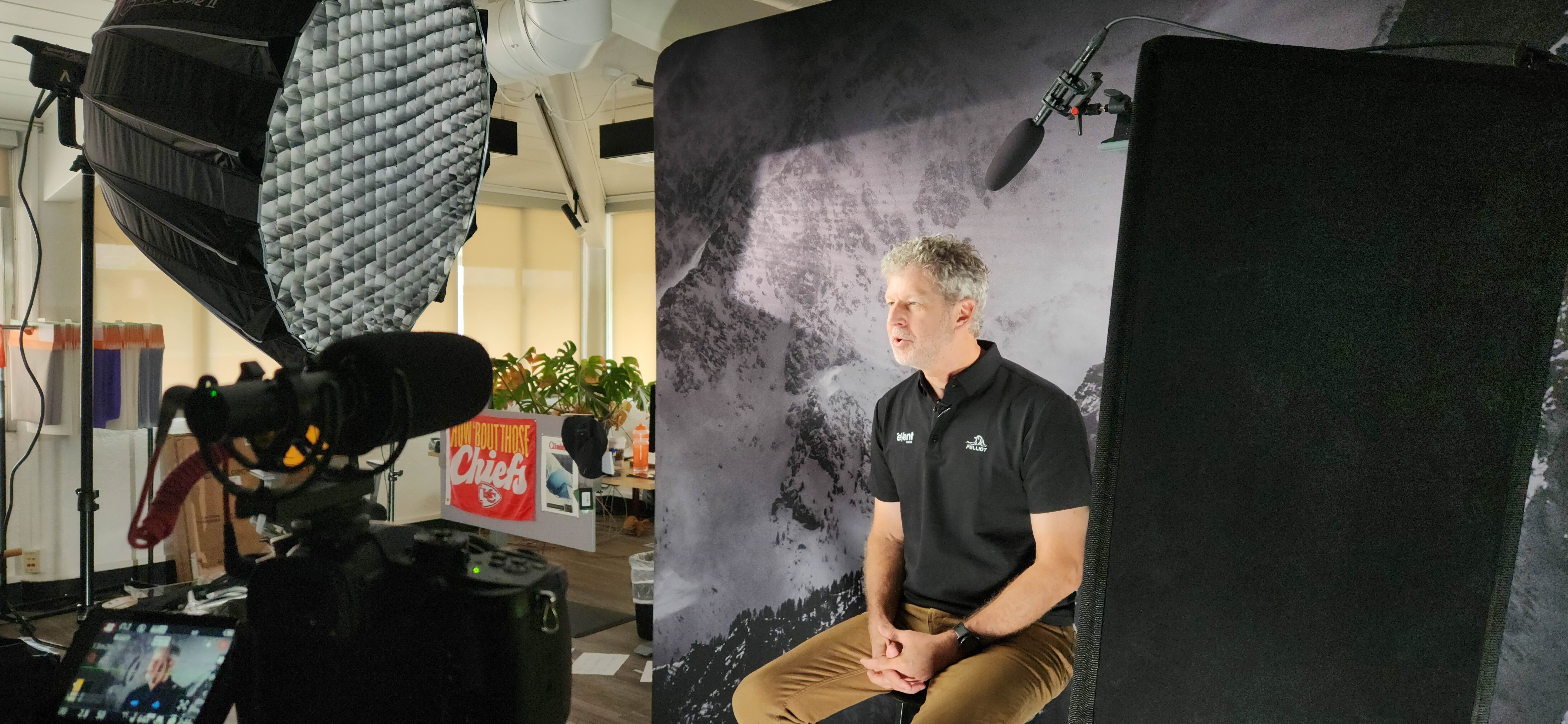 Man in black polo shirt seated and speaking in a video shoot setup with studio lighting and backdrop.