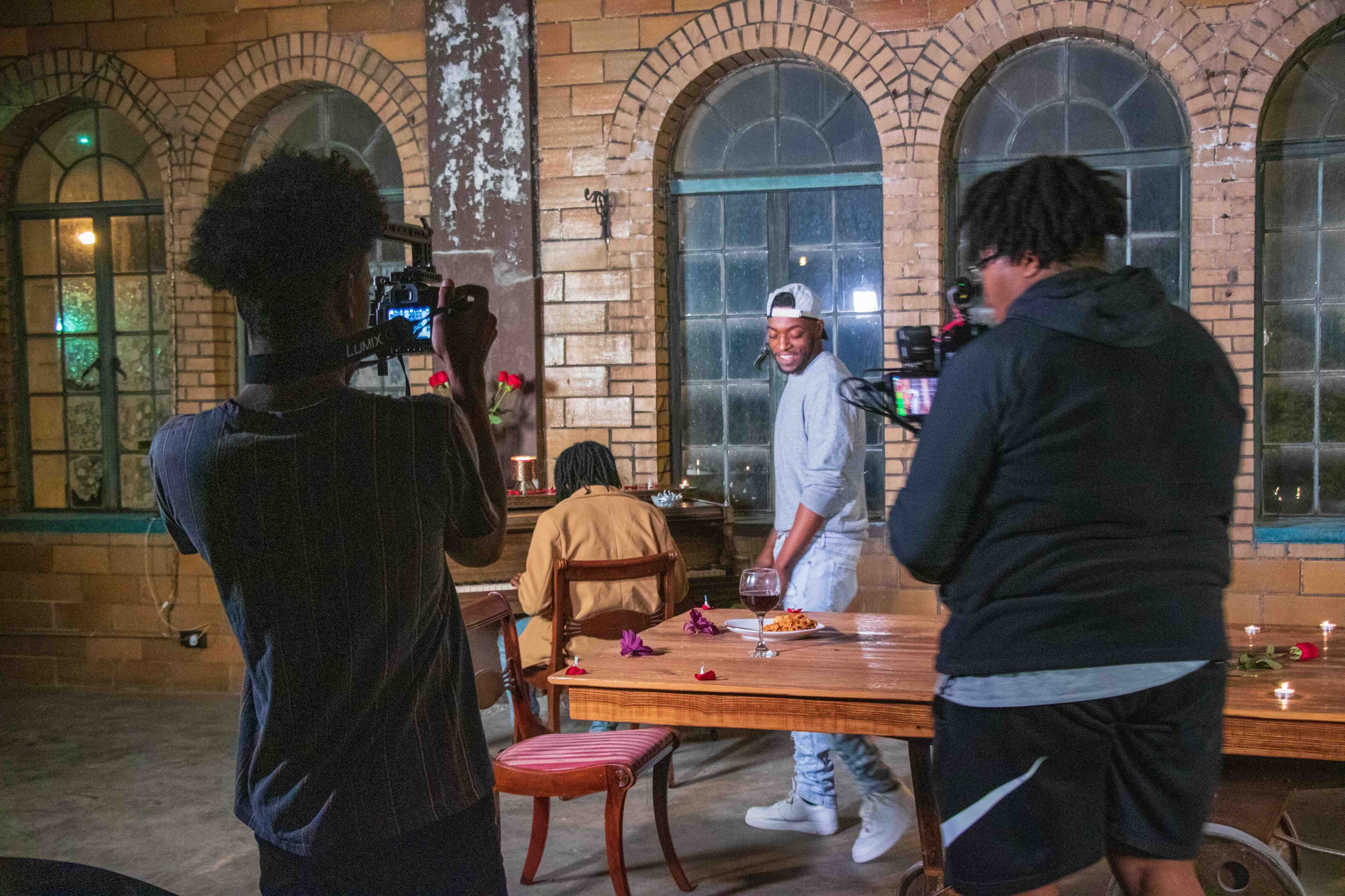 Two men filming another man in a grey shirt standing near a table with food and wine in a rustic room.