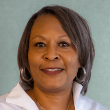 Smiling middle-aged woman with short gray hair and hoop earrings in front of a light blue background.