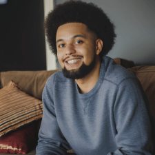 Smiling man with beard and curly hair wearing a gray sweatshirt sitting on a couch.