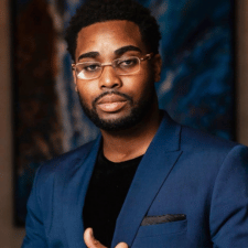 Man with glasses and beard wearing a blue blazer and black shirt against a dark background.