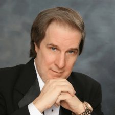 Man in black tuxedo with bow tie resting his chin on folded hands against a gray background.