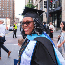 Smiling graduate wearing black cap, gown, and light blue stole standing on city street with people around.