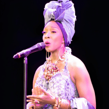 Female singer in a purple outfit and headwrap performing on stage with a microphone. Myself Embodied singer.