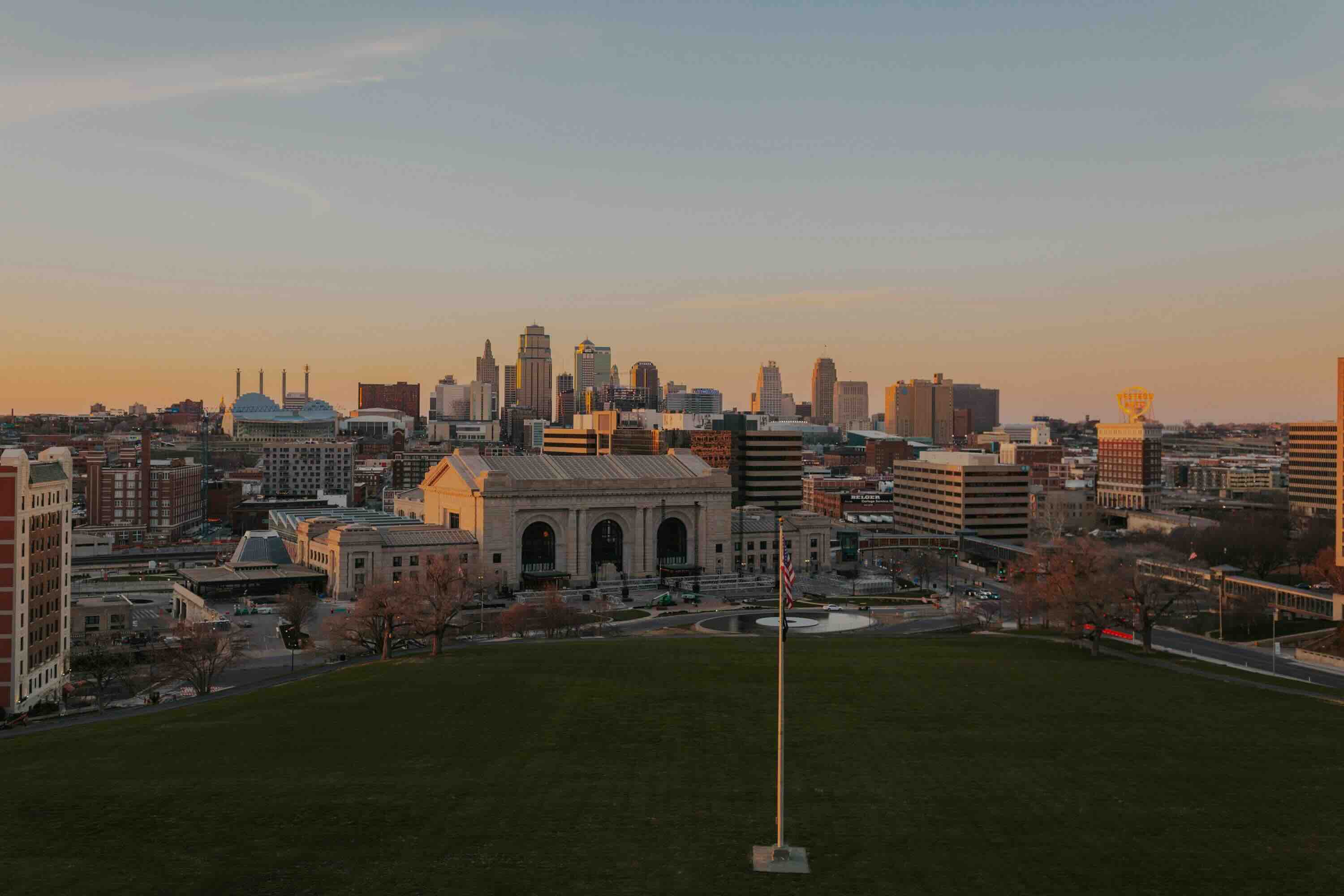 View of Kansas city city skyline at sunset with a green field and flagpole in the foreground.