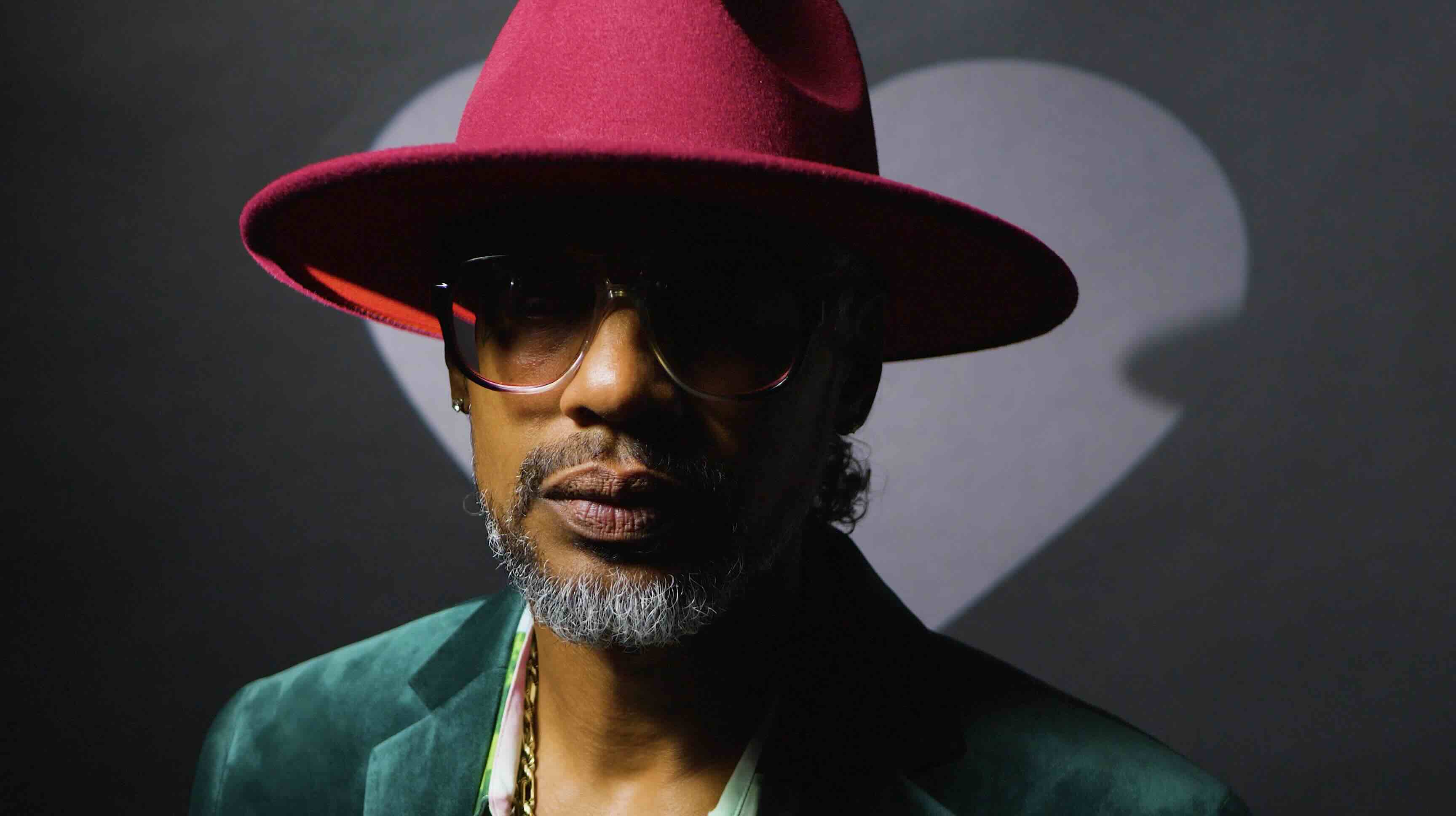 Man with a gray beard wearing a red wide-brimmed hat, sunglasses, green jacket, and gold chain in a music video production shoot.