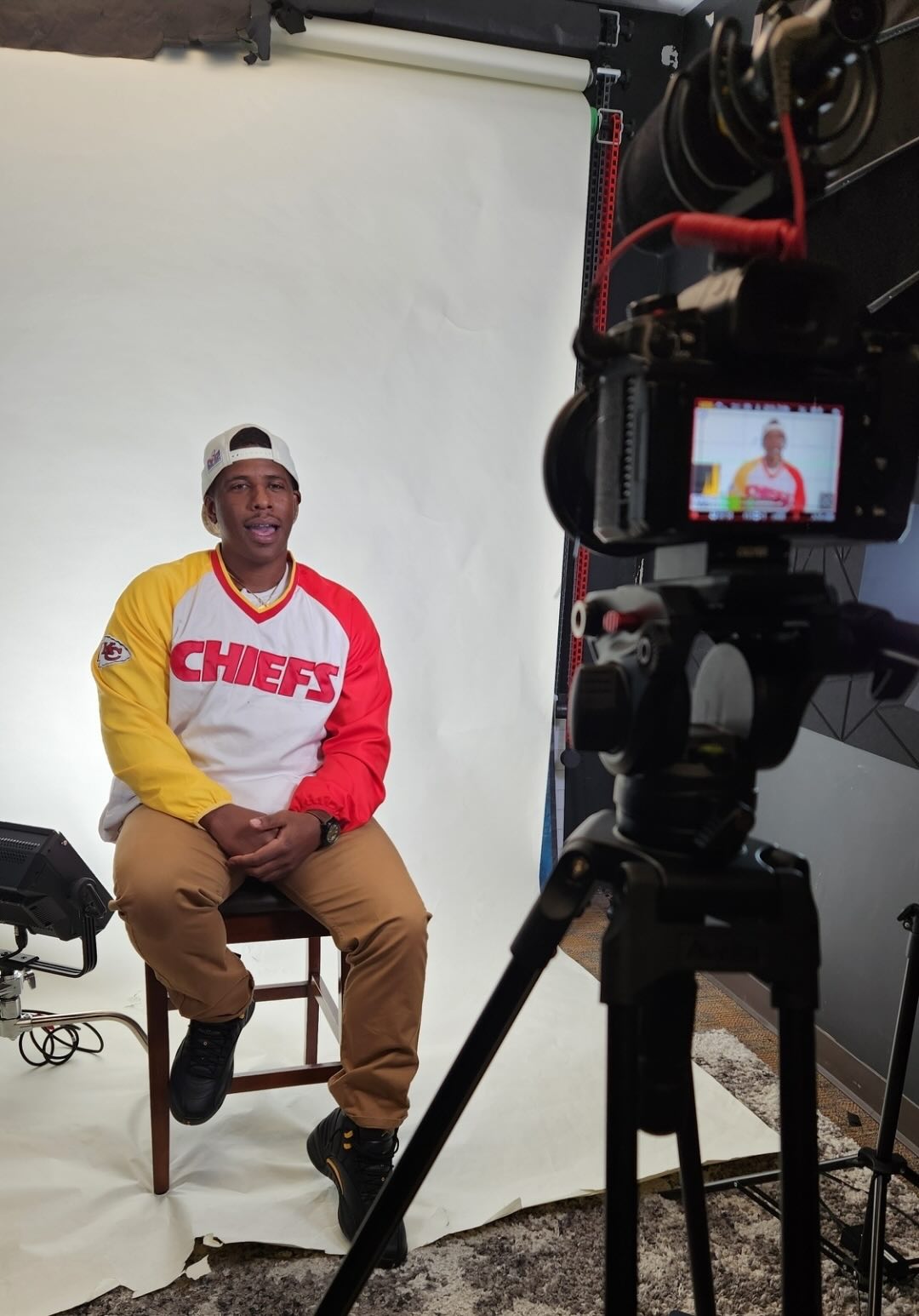Man wearing a Chiefs sweatshirt and white cap, sitting on a chair in front of a camera and white backdrop.
