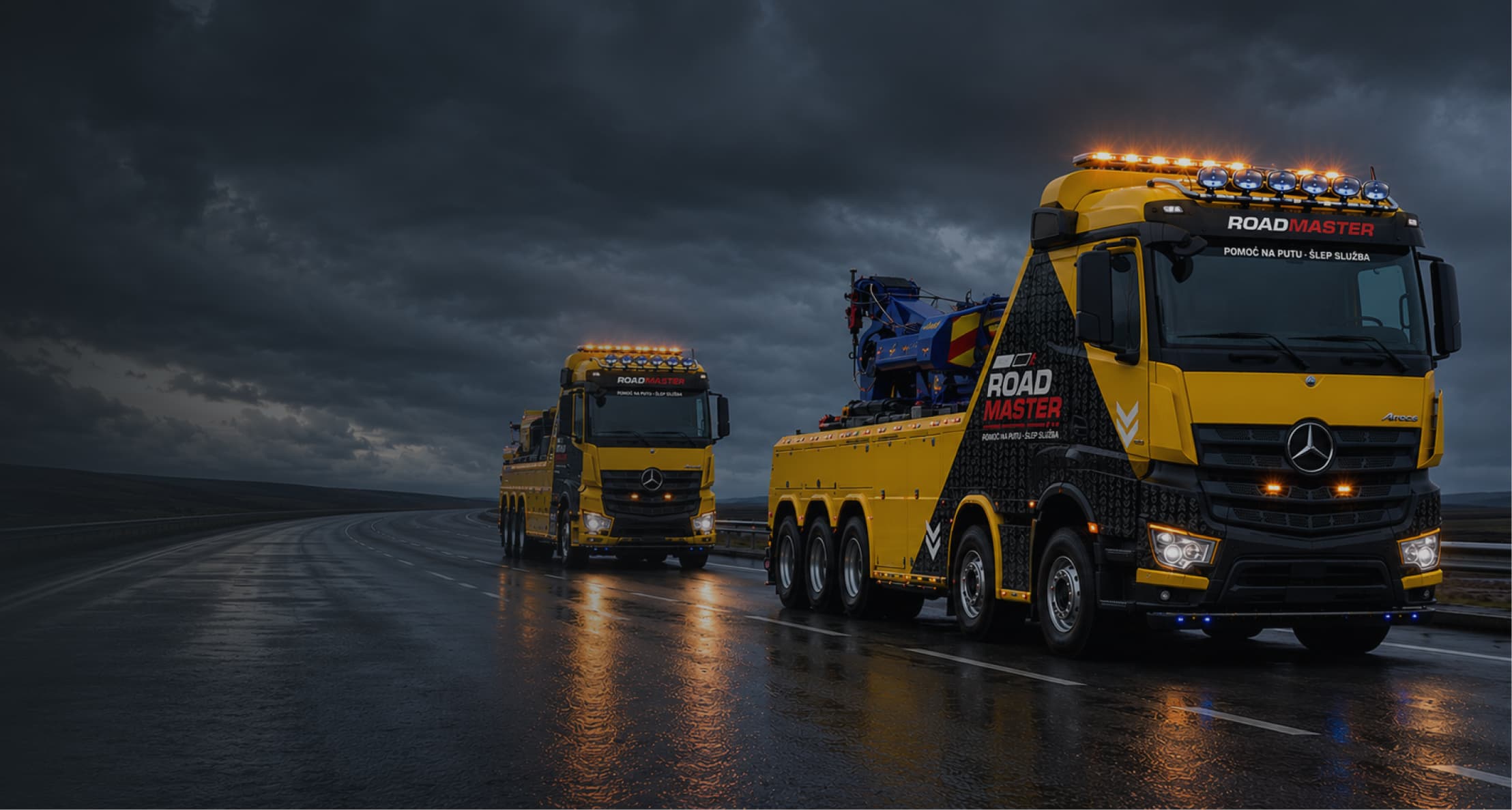 Two bright yellow Road Master tow trucks on a wet highway under dark, cloudy skies at dusk.