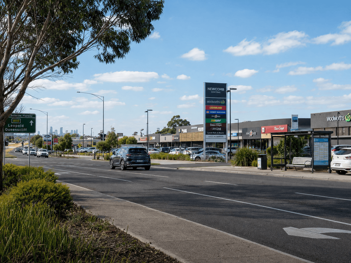 Bellarine Highway passing a modest shopping centre in Newcomb with Geelong in the distance, showing affordable suburb positioning near key amenity and transport.