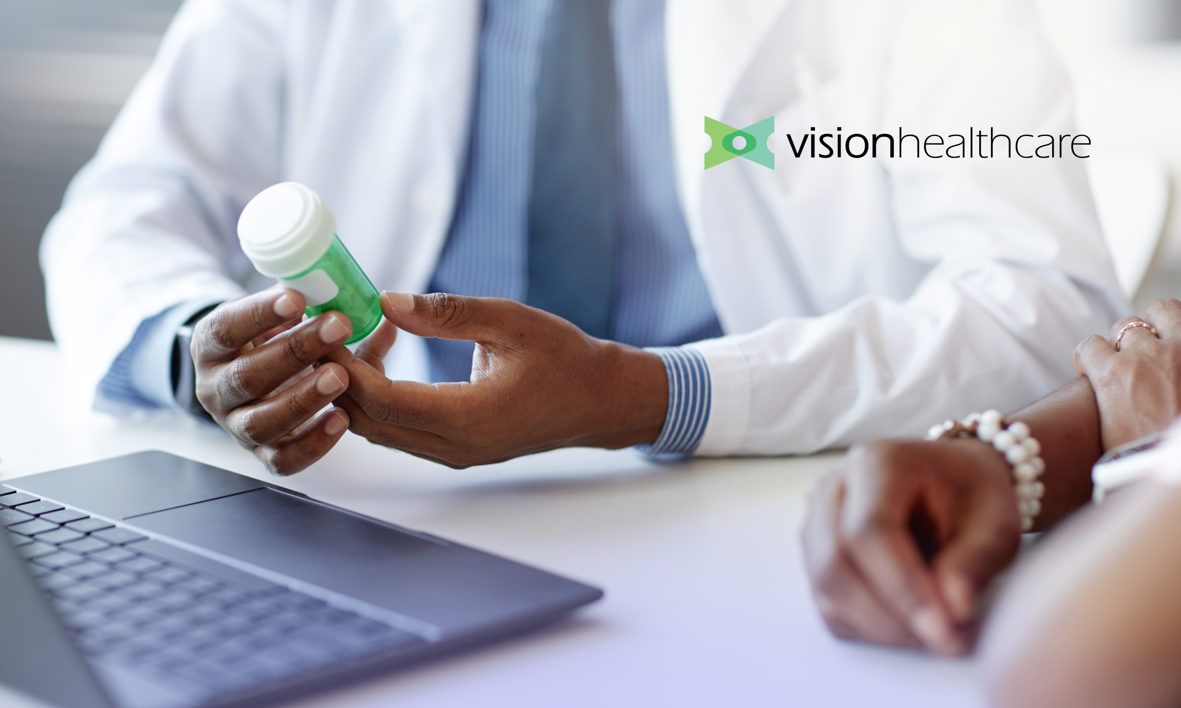 Doctor in white coat holding a green pill bottle while consulting a patient near a laptop.