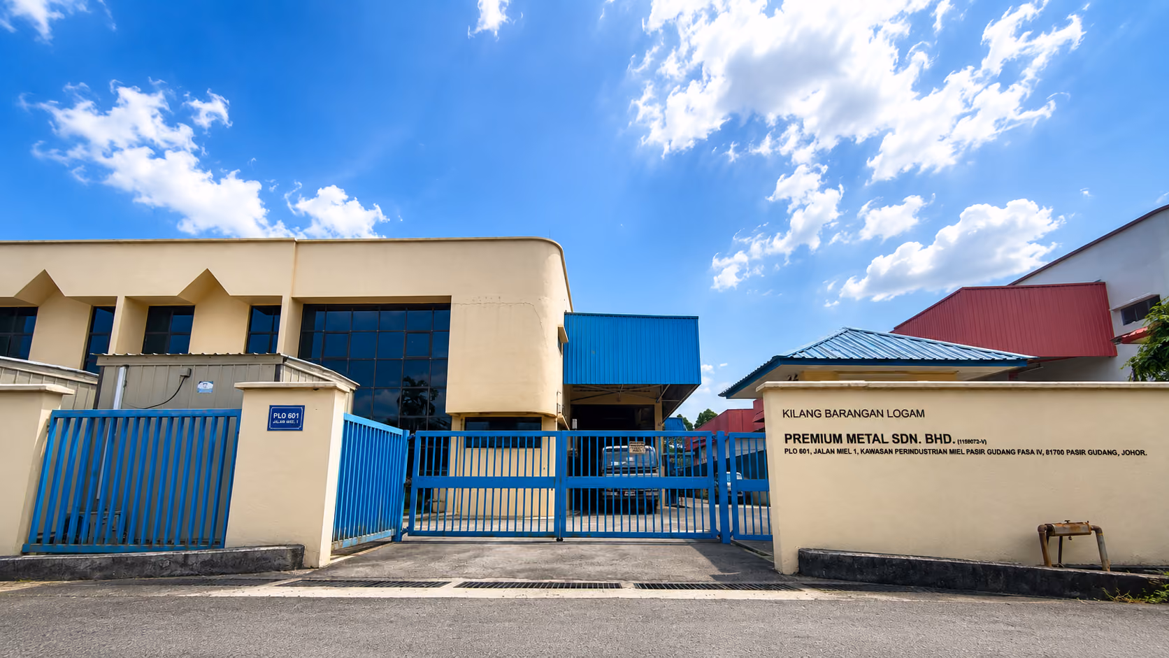 Front view of Premium Metal factory with blue gates under a bright sky with scattered clouds.