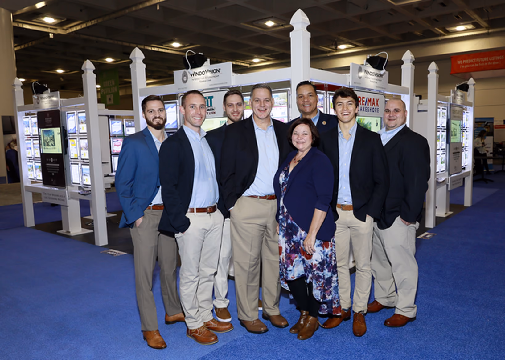 Group of seven men and one woman in business casual attire inside a convention center with display booths.