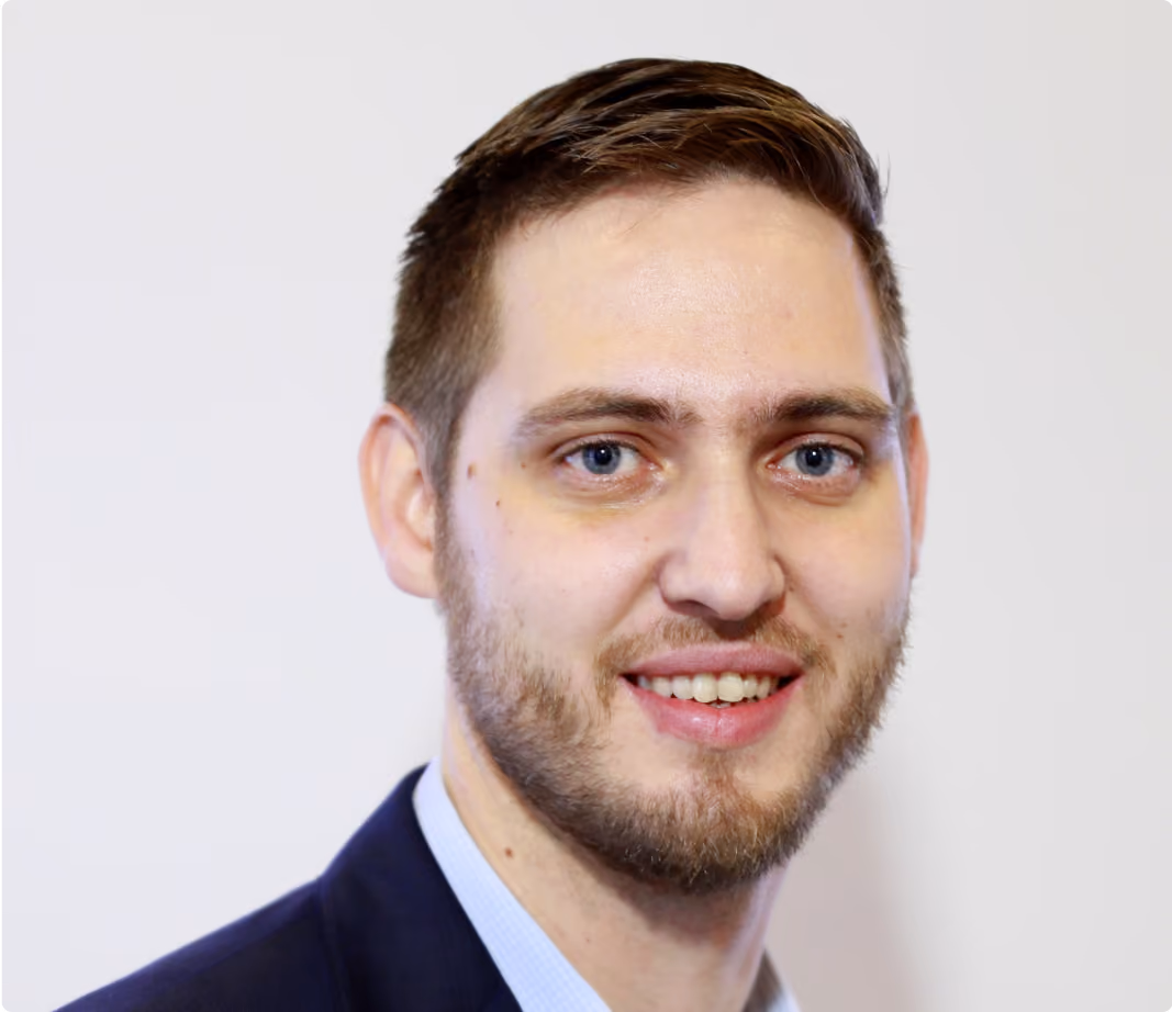 Smiling man with light skin, short brown hair, beard, and blue eyes wearing a dark blazer and light shirt.
