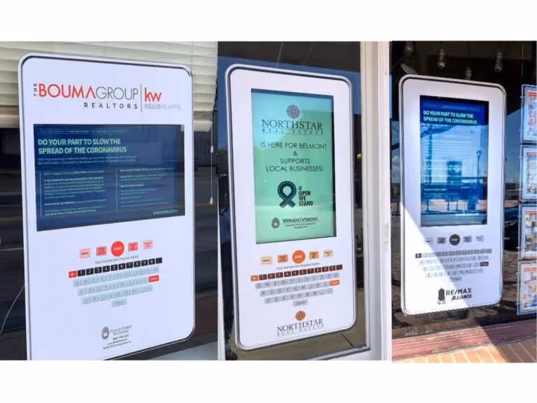 Three real estate digital kiosks displaying public health and local business support messages in storefront windows.