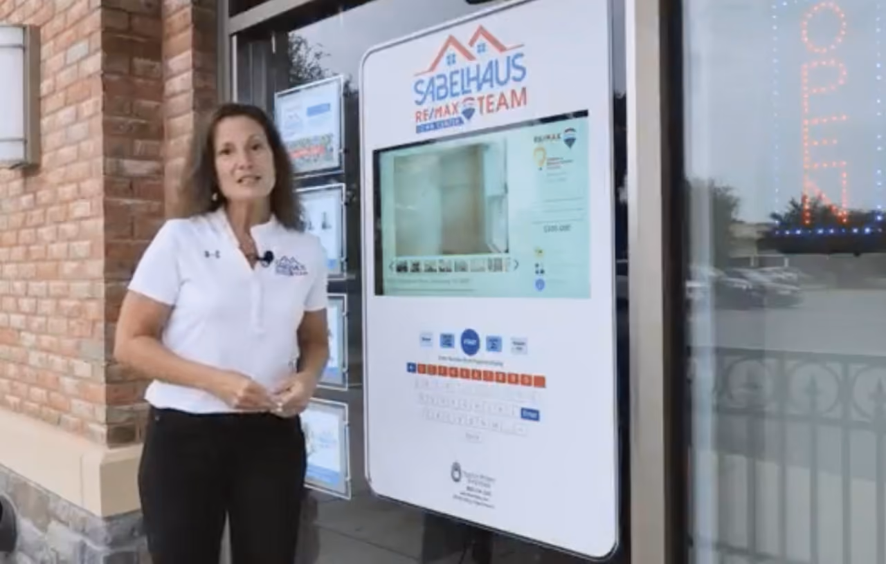 Woman in white shirt stands outside a real estate office near a large digital property display.