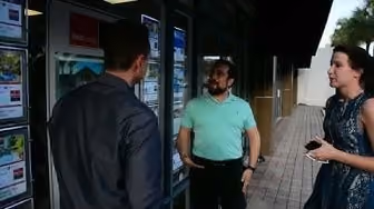 Three people standing outside near a window, two men and a woman talking on a sidewalk.