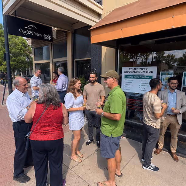 Group of people socializing outside on a sidewalk near storefronts on a sunny day.