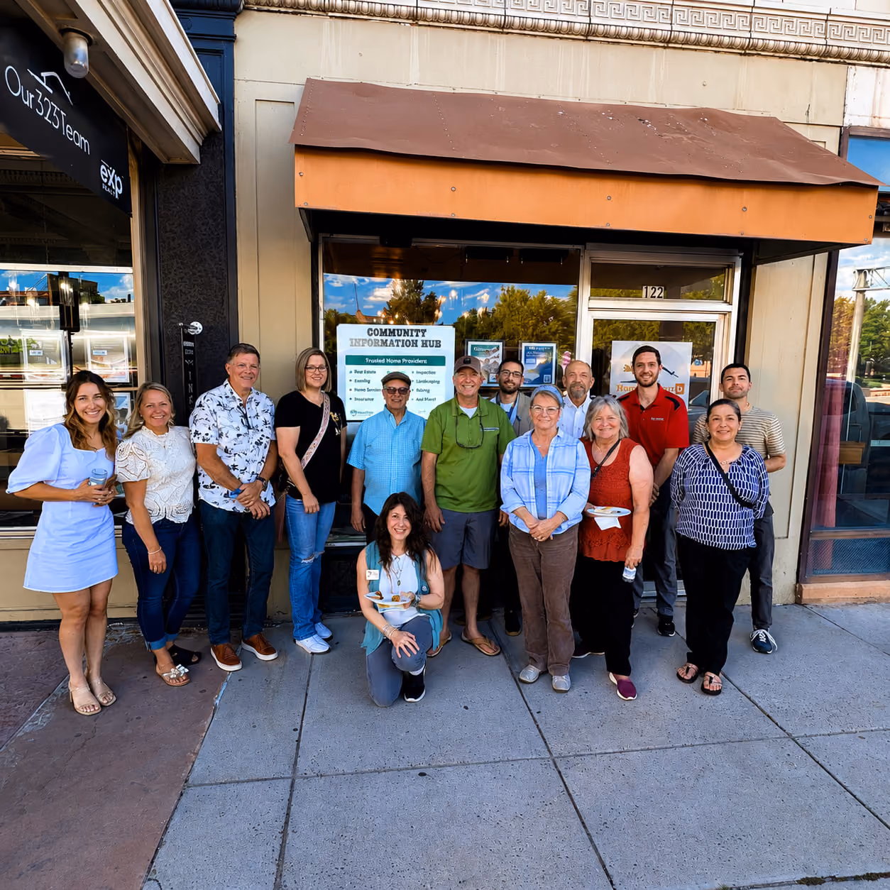 Group of 14 people posing and smiling outside a building under a brown awning.