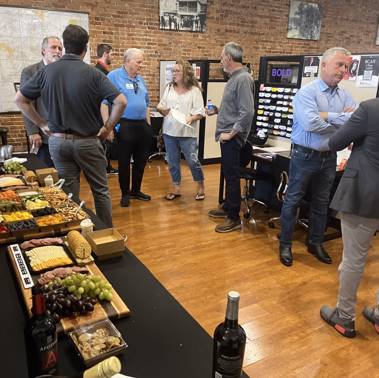 Group of people talking near a table with wine, cheese, fruit, crackers, and cookies in a room with brick walls.