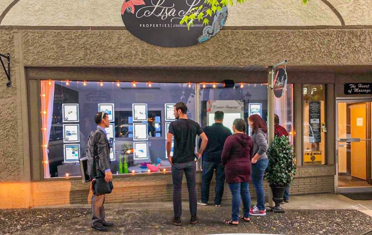 People gathered at a lit real estate office window in the evening, browsing listings