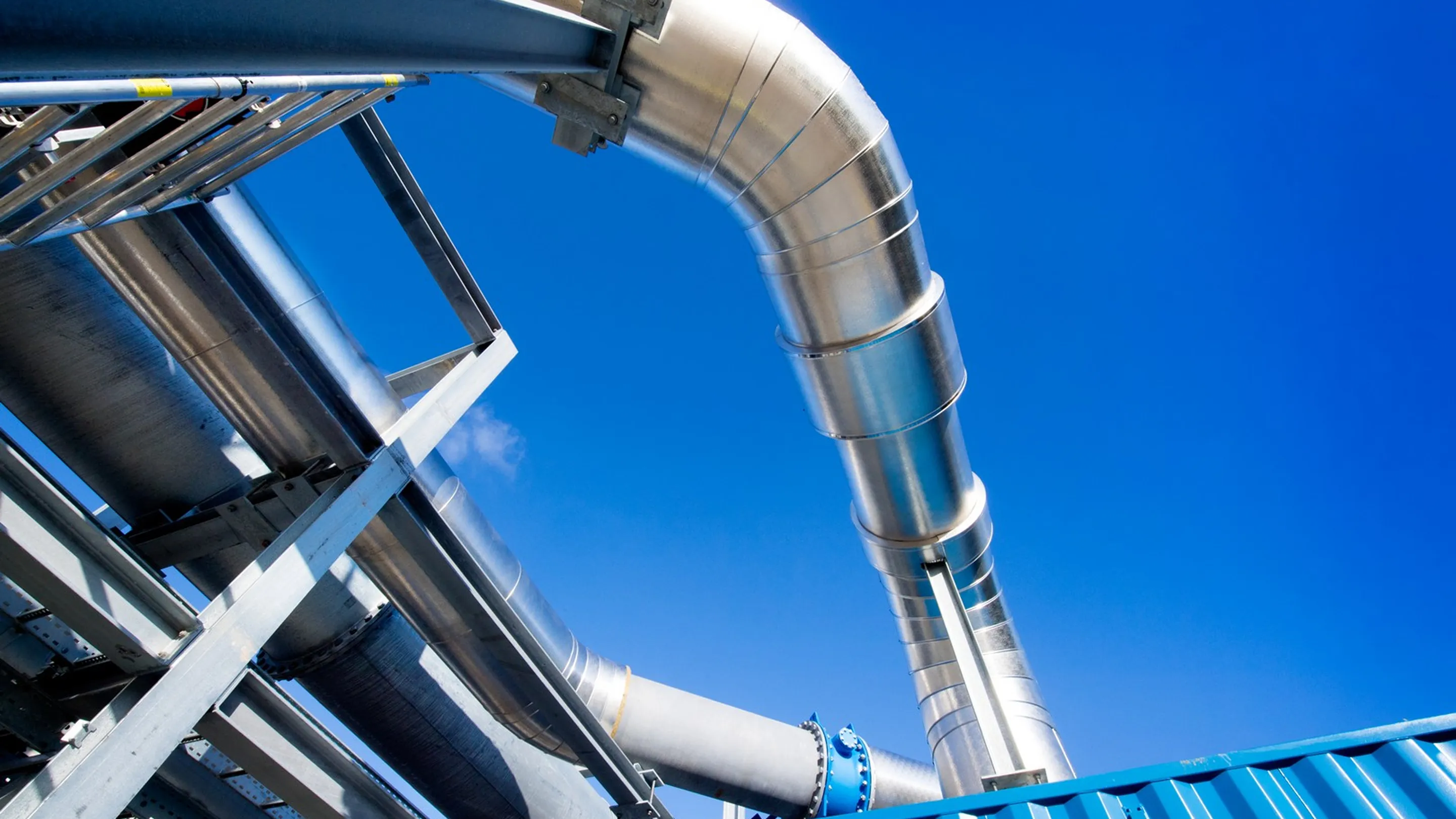 Industriële metalen pijpleiding met gebogen buis en blauwe bevestigingen tegen een helderblauwe lucht.