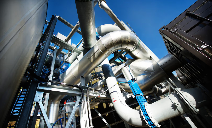 Industriële installatie met grote metalen pijpleidingen en buizen onder een heldere blauwe lucht.