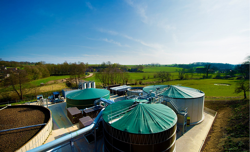 Luchtfoto van een biovergistingsinstallatie in het groen, met grote ronde tanks met groene dekzeilen en pijpleidingen onder een heldere blauwe lucht.