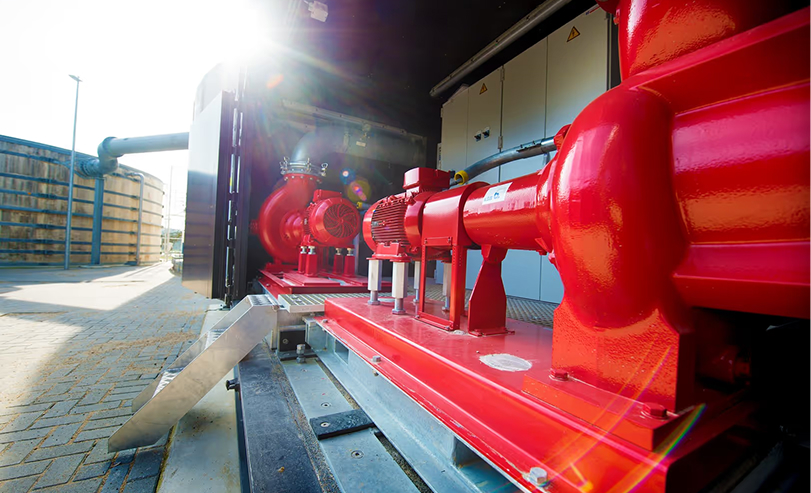 Rode industriële pompinstallatie binnen een technische ruimte met zonlicht dat naar binnen schijnt en een pijpleiding buiten zichtbaar op een bestrate ondergrond.