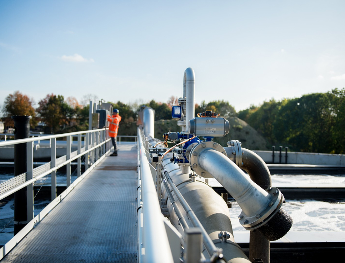 Binnenzicht van een industrieel waterzuiveringsstation met metalen leidingen en apparatuur, en een persoon in een oranje veiligheidsjas die aan een bedieningspaneel werkt vanwege een hekwerk, met groene bomen op de achtergrond onder een lichtbewolkte hemel.