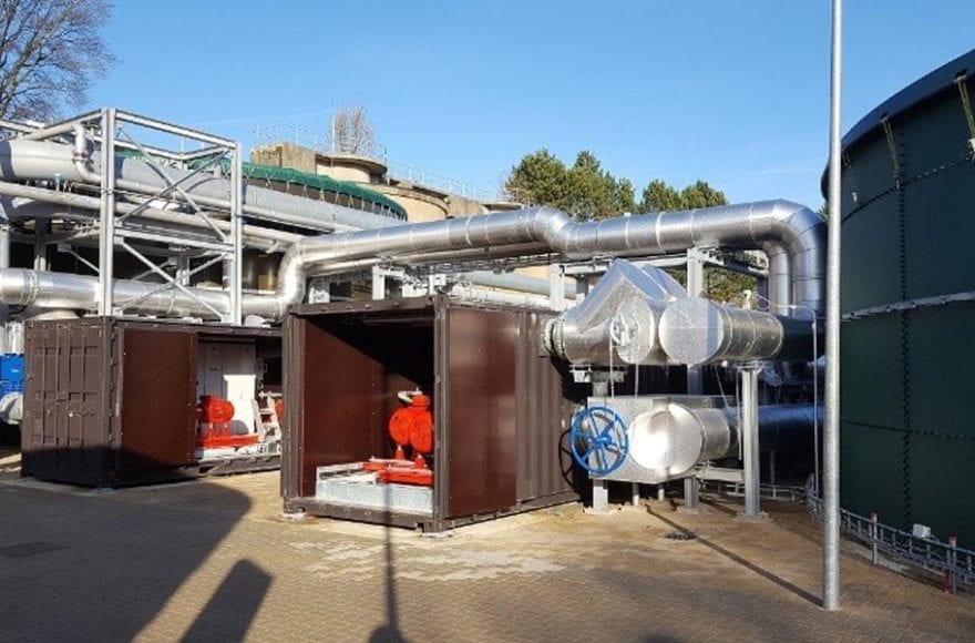 Industrieel terrein met grote metalen pijpleidingen en twee open containers met rode machines binnenin, onder een heldere blauwe lucht.