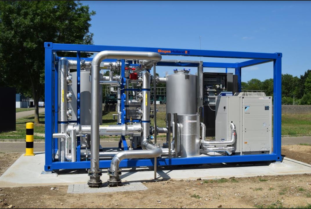 Blauwe metalen frame met roestvrijstalen leidingen en apparatuur buiten op een betonnen platform, onder een heldere blauwe lucht met bomen op de achtergrond.