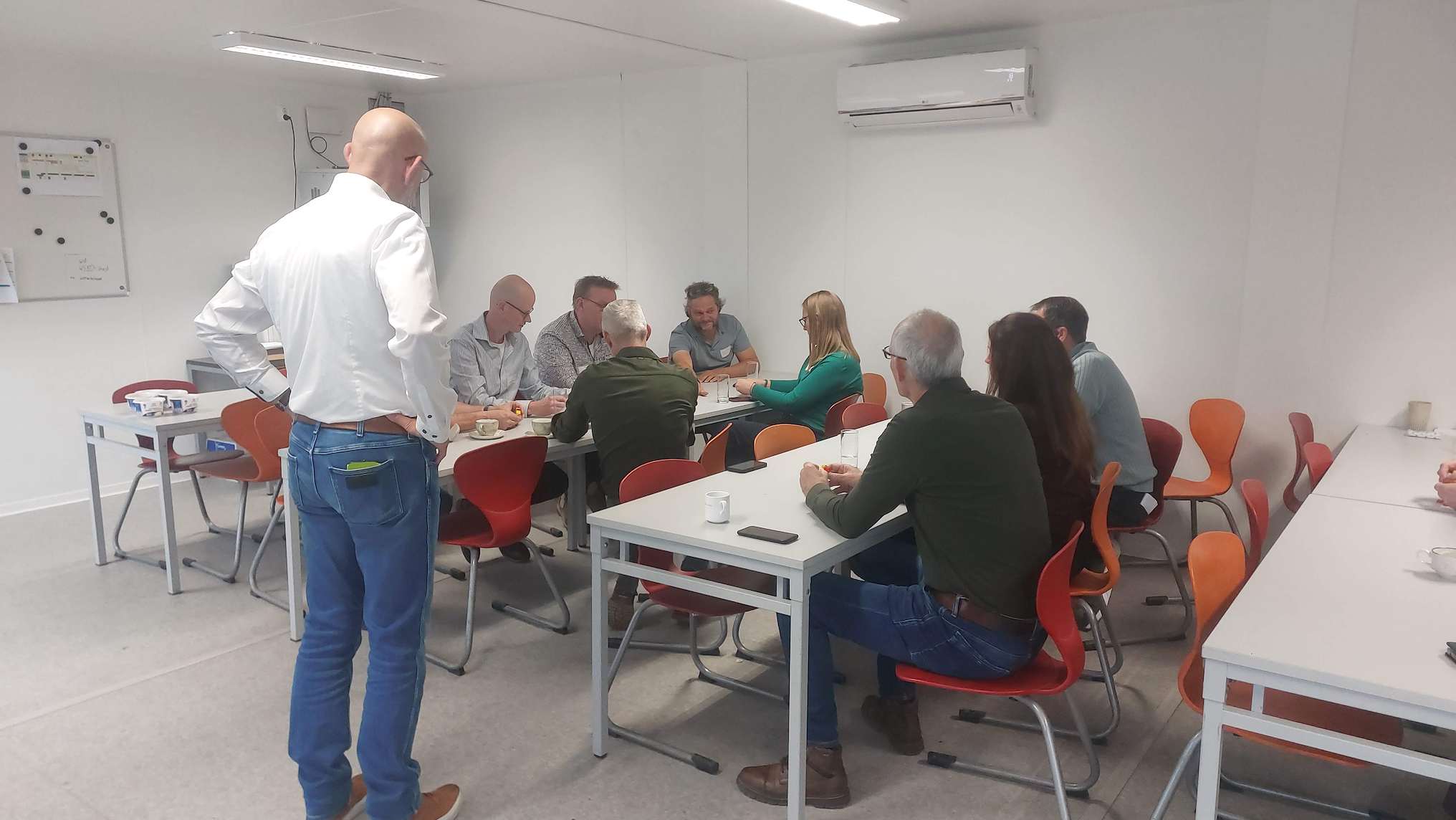 Een groep mensen zit rond tafels in een eenvoudige vergaderruimte met witte muren, een airco en oranje, rode stoelen, terwijl een staande man in een witte overhemd en blauwe jeans hen observeert.