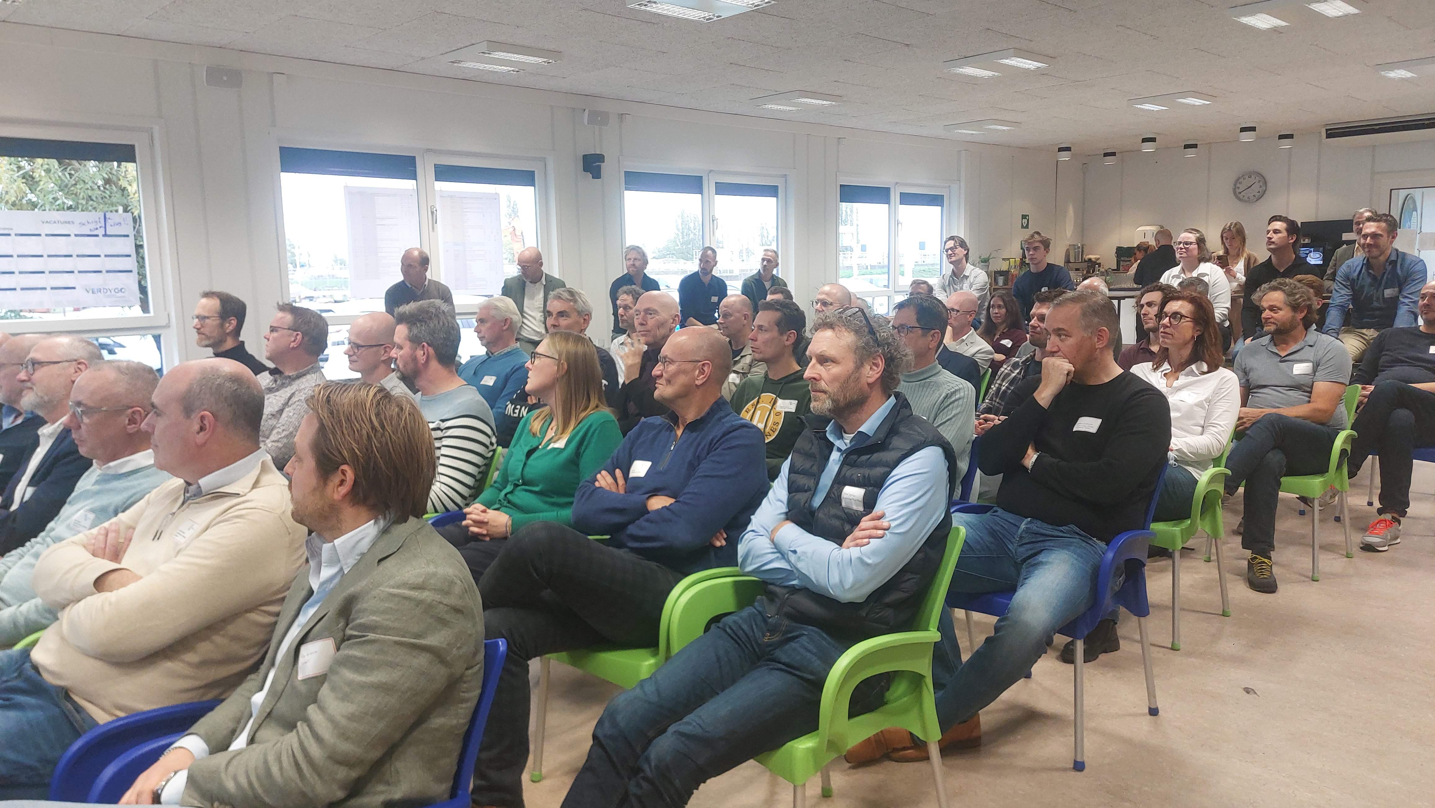 Een groep volwassenen zit in een goed verlichte kamer op kleurrijke stoelen en kijkt aandachtig naar voren, mogelijk tijdens een presentatie of lezing.