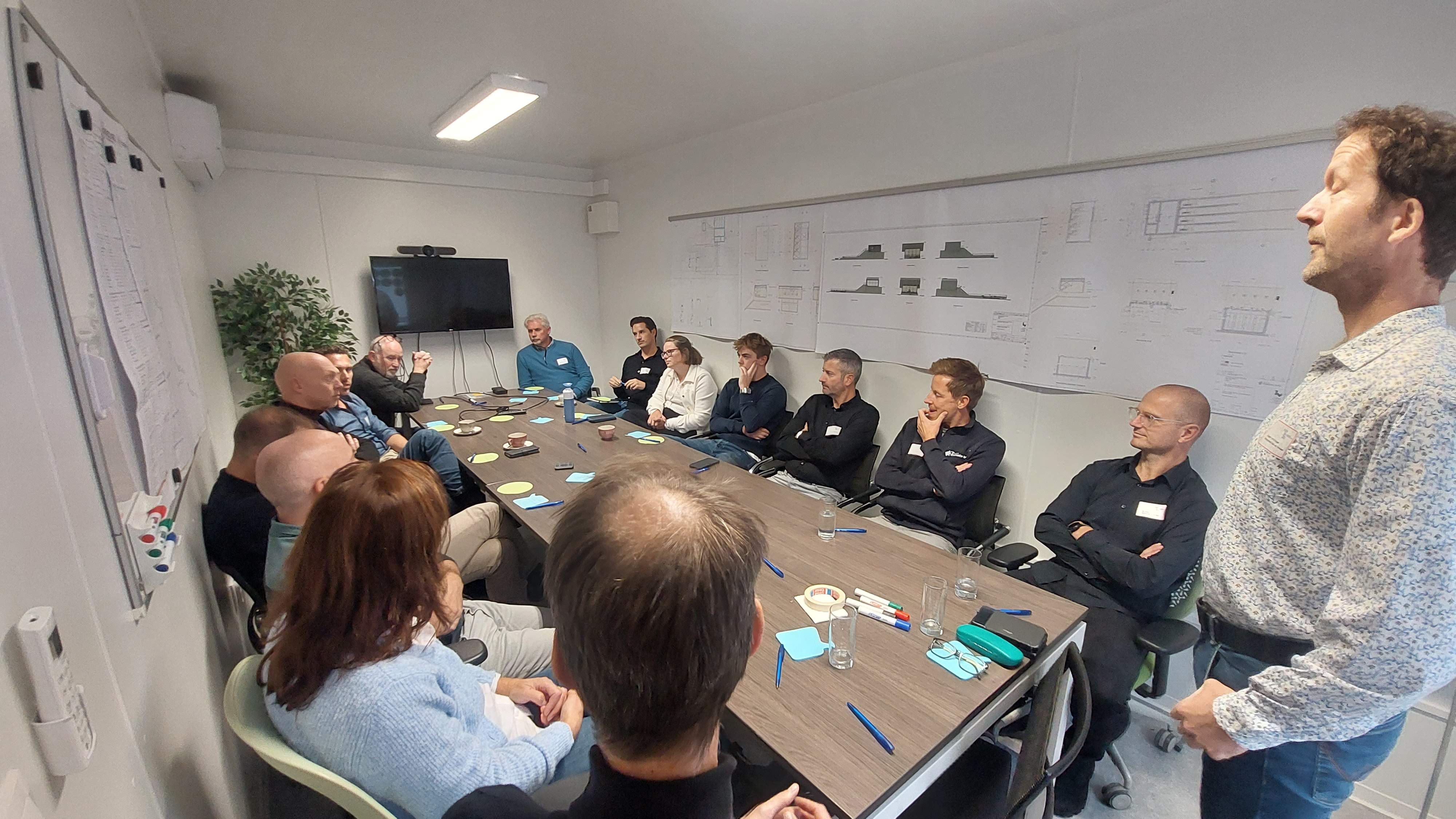 Een groep van vijftien mensen zit rond een grote conferentietafel in een vergaderruimte. Een man staat aan de rechterkant en spreekt waarschijnlijk de groep toe. Aan de muur hangen meerdere technische tekeningen. Op de tafel liggen pennen, memoblaadjes, bekers en enkele apparaten. De ruimte is neutraal ingericht met een tv-scherm aan de achterwand en een plantenpot in de hoek.