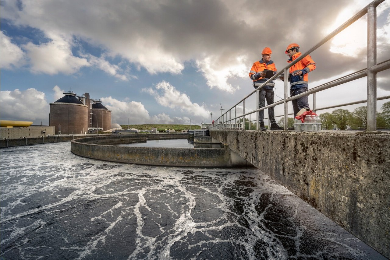 Twee werknemers in oranje veiligheidskleding en helmen bespreken iets op een tablet bij een waterzuiveringsinstallatie met schuimend water en grote opslagtanks onder een bewolkte hemel.